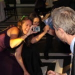 Two women bent down with a camera pointed at a man holding a drink, dancing on the dance floor with Paul Michaels Events DJ Company playing music