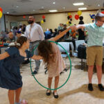 A group of kids holding hands in a chain link and passing a hula hoop to each other using their bodies, with entertainment being provided by Paul Michaels Events, an expert in B'nai Mitzvah entertainment services